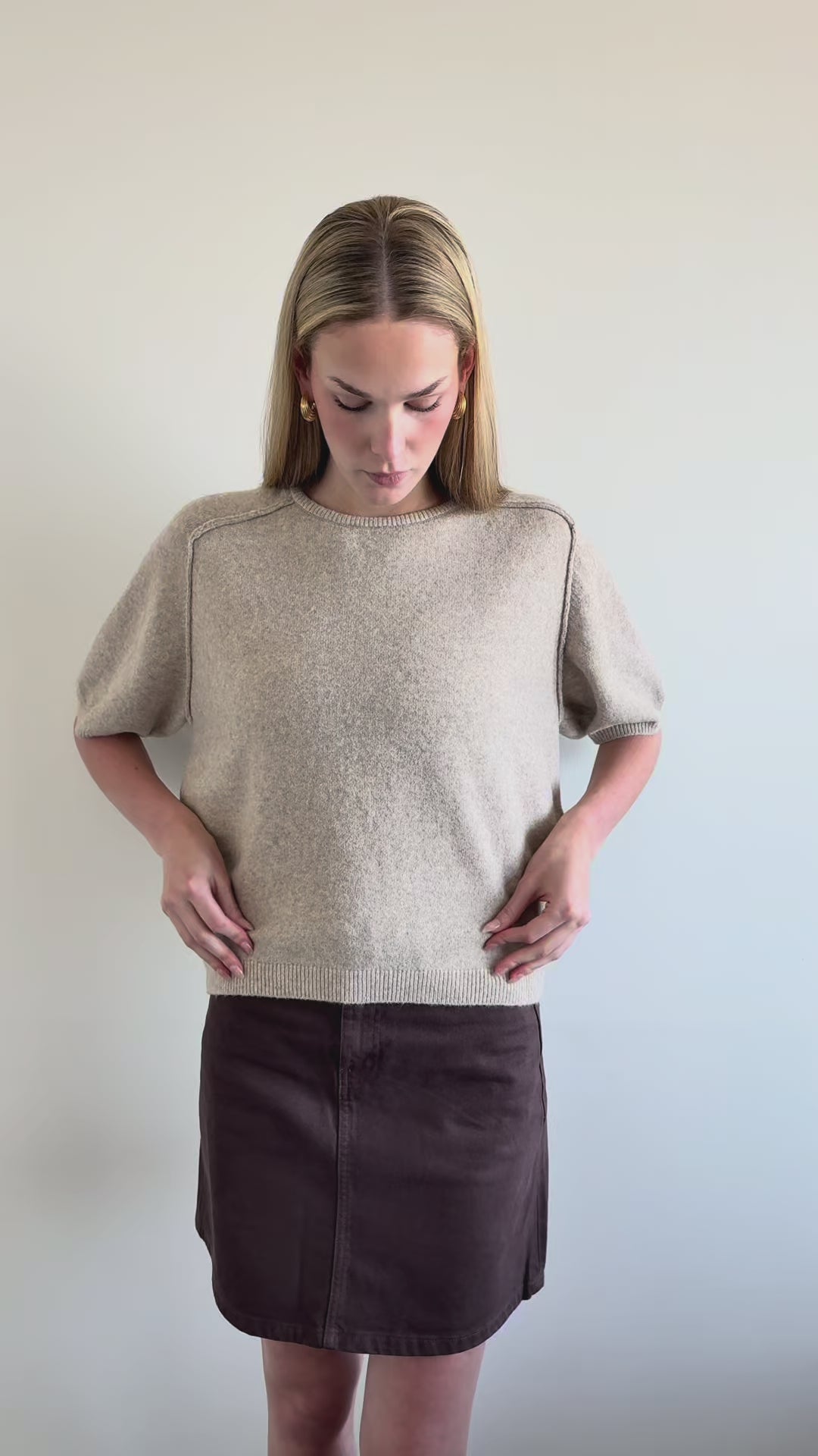 A woman modeling a light beige short-sleeve knit top and a dark brown mini skirt with a belt, standing against a neutral textured fabric backdrop.