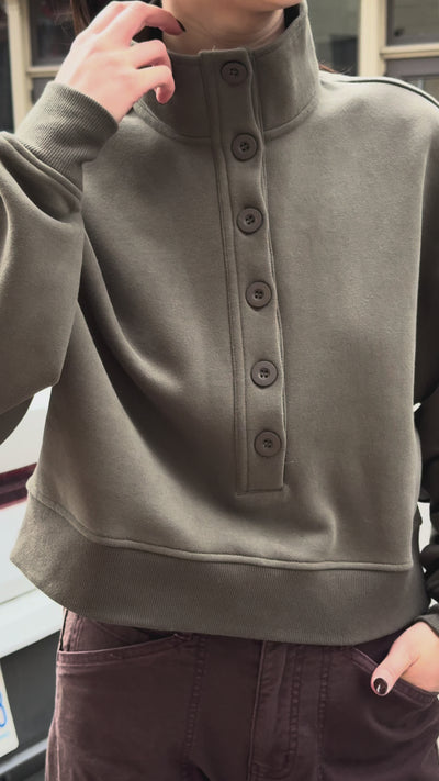 A woman wearing an olive green cropped button-front sweatshirt and dark brown pants, posing outdoors against a stone building background.