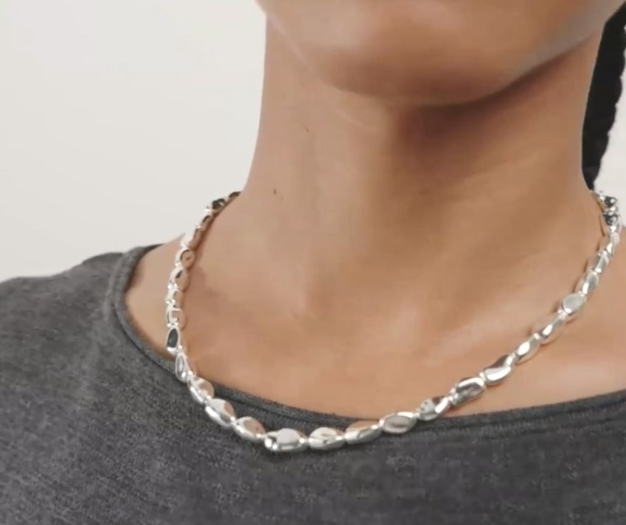 A close-up of a model wearing a chunky silver chain necklace with irregular, polished links over a black crewneck shirt.
