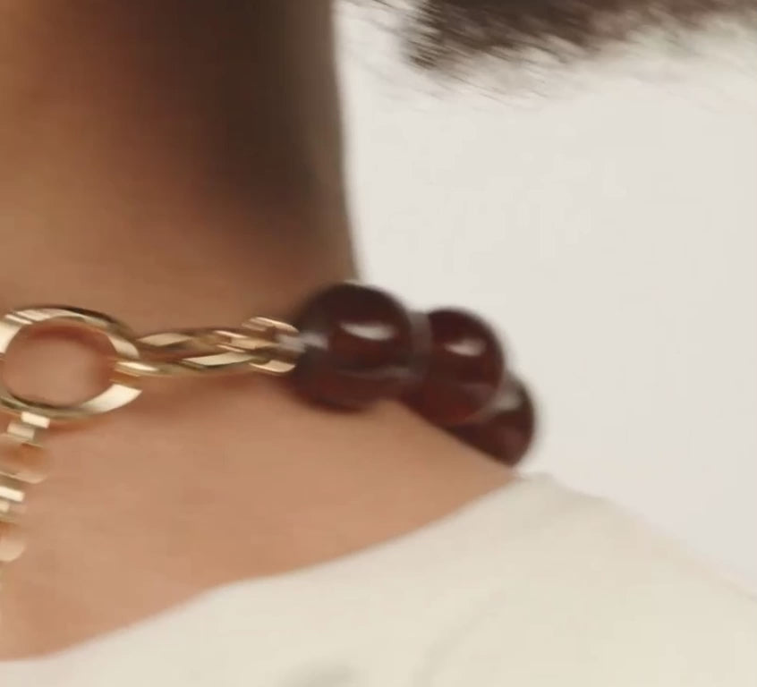 A close-up of a model wearing a dark reddish-brown beaded necklace with polished round beads, paired with a white crew-neck top against a light gray background.