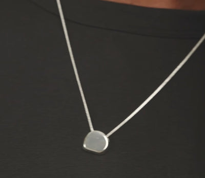 Two models wearing minimalist silver pendant necklaces with flat, geometric oval pendants against a light gray background.