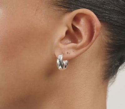 A close-up side profile of a woman with dark curly hair wearing a small silver hoop earring with a black and white patterned center, against a light gray background.