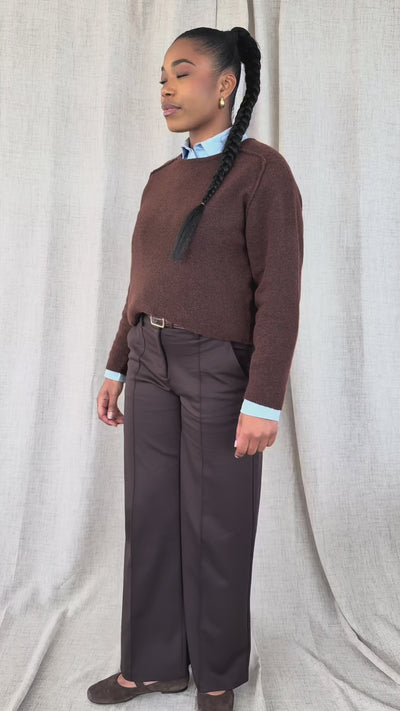 A woman wearing a brown knit crewneck sweater and dark gray trousers, standing against a neutral beige fabric backdrop.
