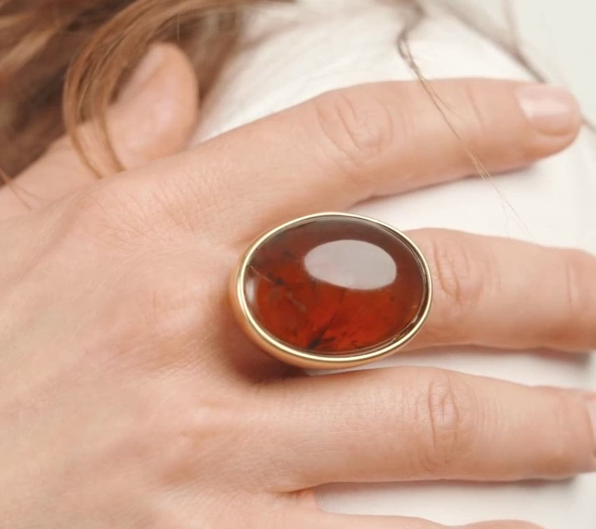 A gold ring with a large oval dark brown gemstone, worn on a person's finger against a clean white background.