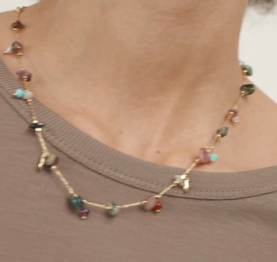 A close-up of a model wearing a delicate gold chain necklace adorned with small, colorful, irregularly shaped gemstone beads. The model is wearing a ribbed brown top against a clean, light off-white background.