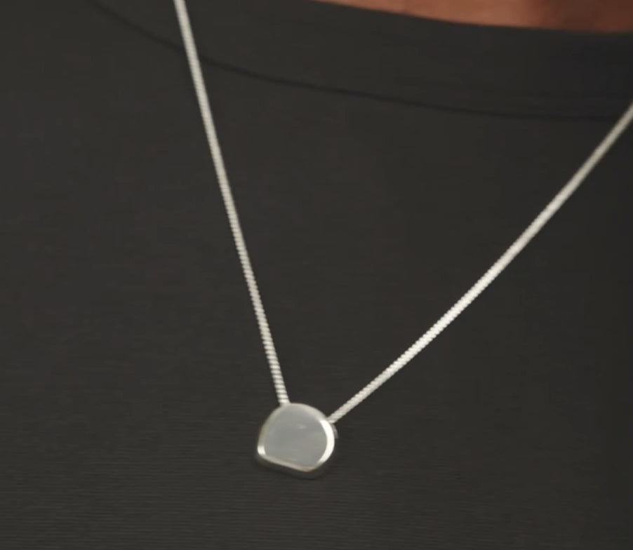 A close-up of a model wearing a delicate gold chain necklace with a small, flat, circular gold pendant. The model is wearing a black ribbed tank top, and her curly dark hair is visible against a light background.