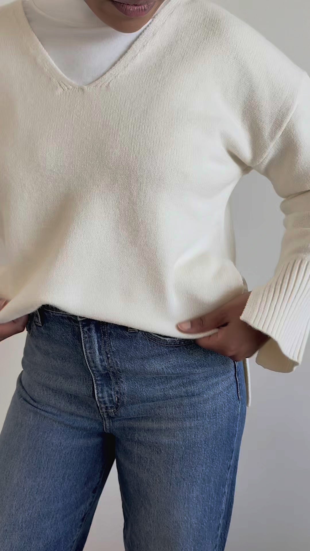 A woman with a high braided ponytail is shown from the side, wearing a cream-colored knit sweater layered over a white mock-neck top and blue jeans, standing against a neutral beige fabric backdrop.