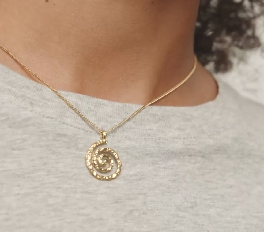 A gold spiral pendant necklace worn by a model in a light gray heathered t-shirt.