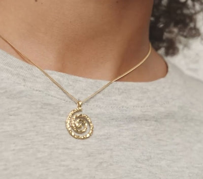 A gold spiral pendant necklace worn by a model in a light gray heathered t-shirt.
