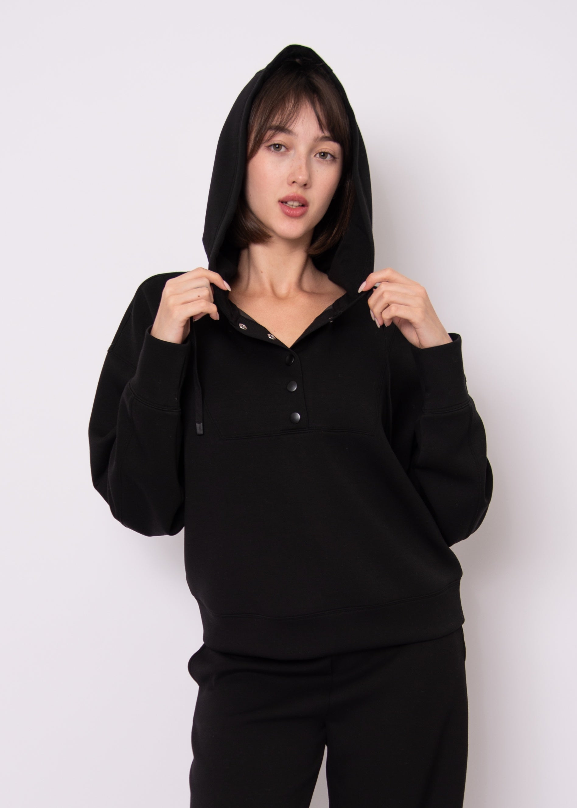A woman wearing a black pullover hoodie with a three-button placket and matching black pants, standing against a plain light gray studio background.