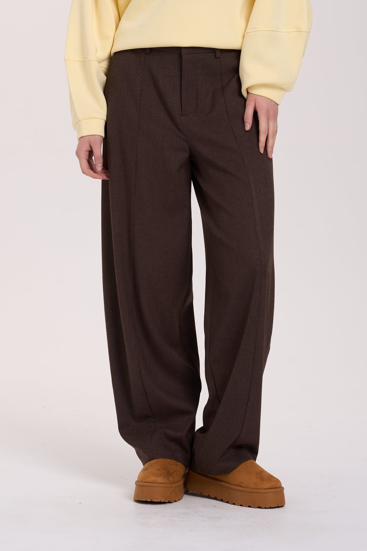 A person wearing dark brown wide-leg pleated trousers, styled with a pale yellow sweatshirt and brown platform slippers against a plain light background.