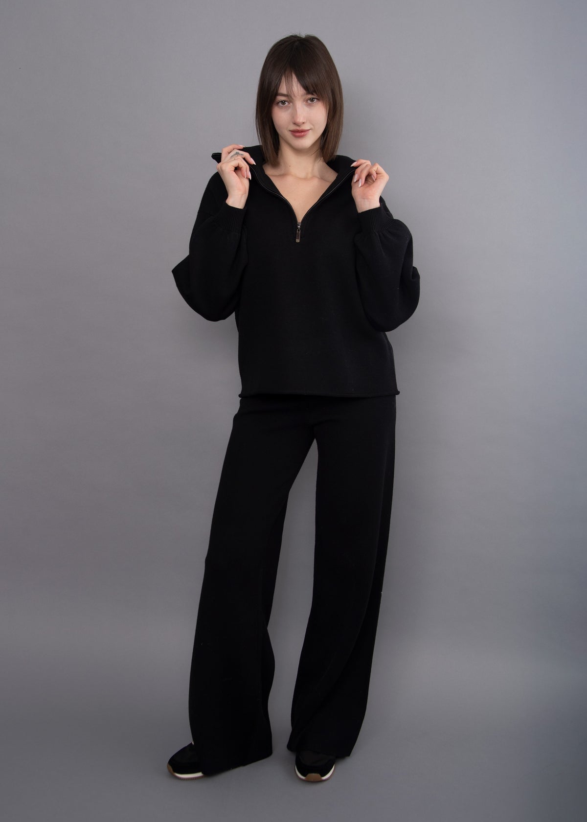 A woman modeling a black two-piece loungewear set consisting of a half-zip hoodie and matching wide-leg pants, standing against a solid gray studio background.