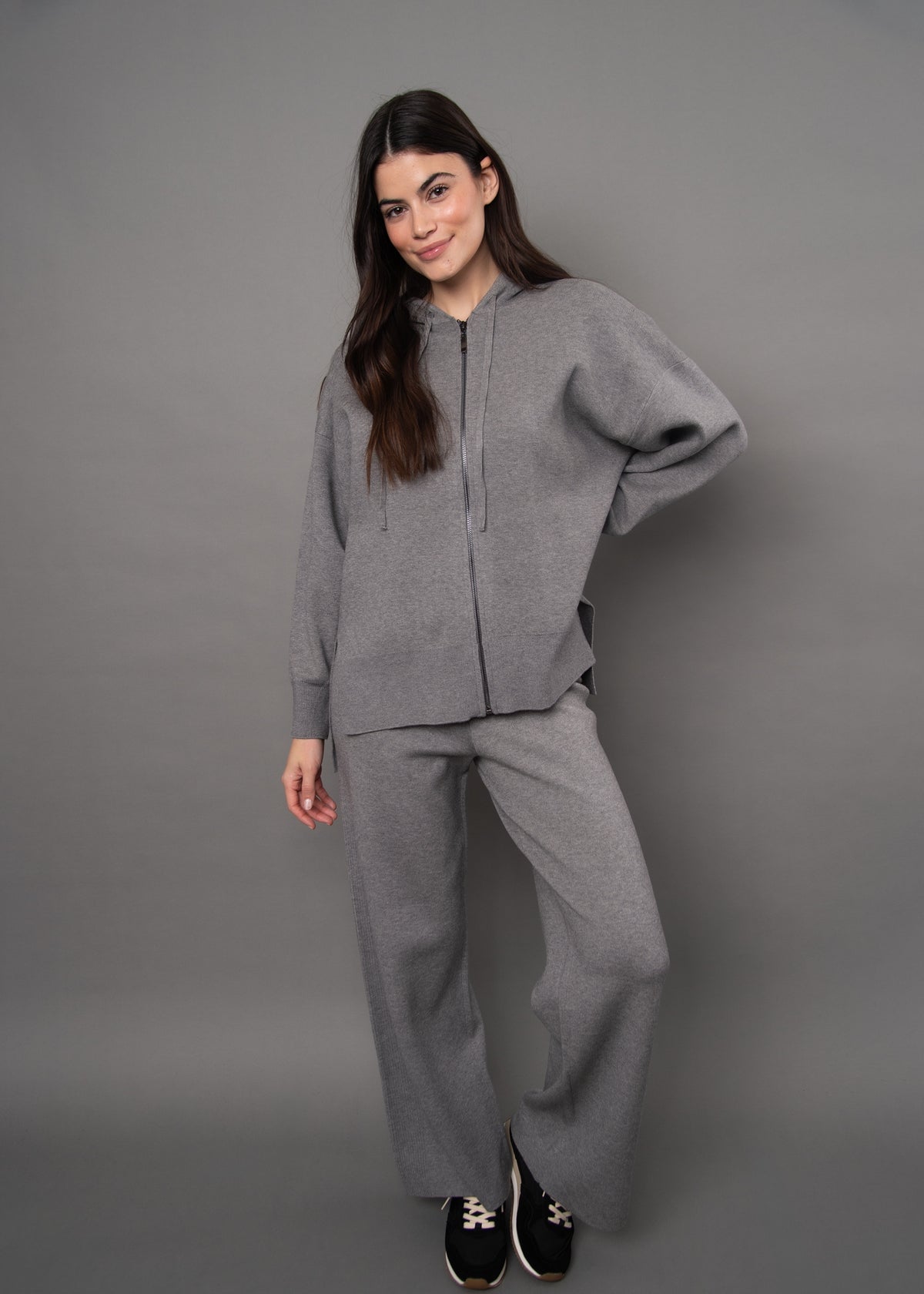 A woman modeling a matching gray knit lounge set consisting of a cropped zip-front hoodie and wide-leg sweatpants, paired with black sneakers, against a solid gray studio background.